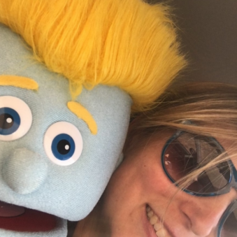 Tiffiny Clifton poses with a blue muppet with yellow hair and eyebrows. Tiffiny is wearing big sunglasses.