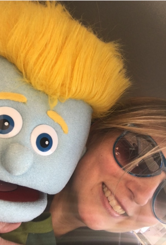 Tiffiny Clifton poses with a blue muppet with yellow hair and eyebrows. Tiffiny is wearing big sunglasses.